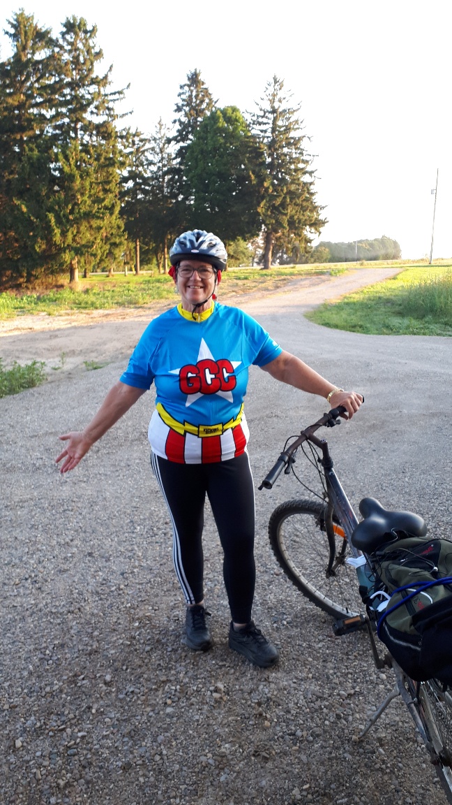Woman in bicycle helmet and bicycle jersey with a Superhereo's star on the front, is smiling with one arm outstretched and the other holding her bike