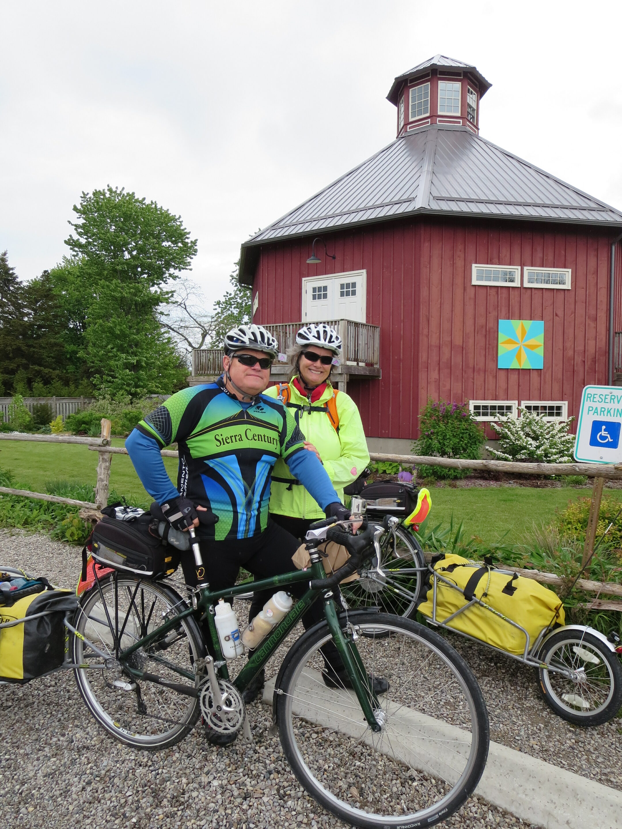 A man and a woman wearing cycling clothes and helmets stand with their bicycles laden with packs in front of the a red eight sided barn
